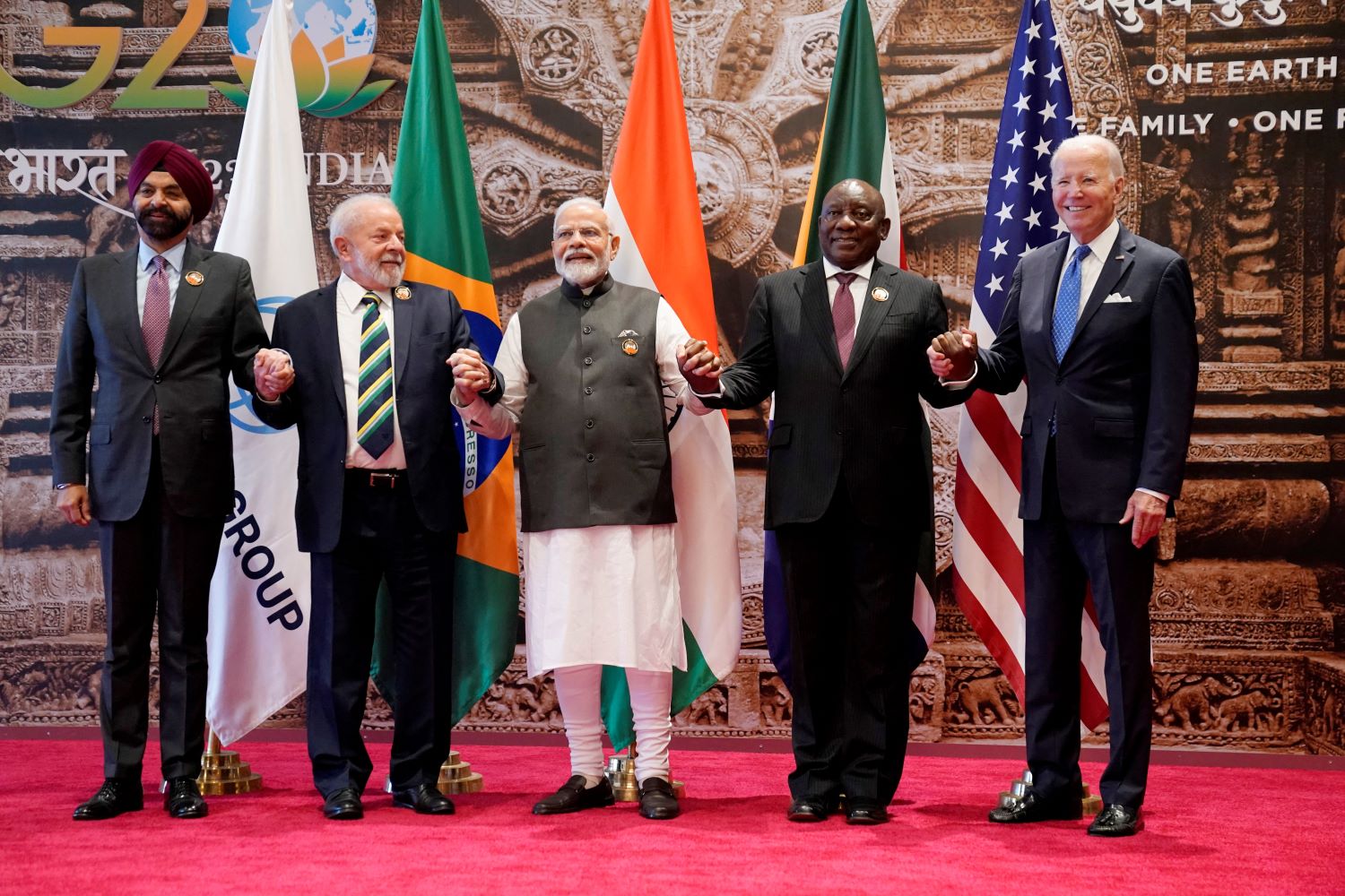 World Bank President Ajay Banga, Brazilian President Luiz Inacio Lula da Silva, Indian Prime Minister Narendra Modi, South African President Cyril Ramaphosa, and U.S. President Joe Biden hold hands on a red carpet with representative flags behind them, posing for a group photo during G20 Summit in New Delhi, India on Saturday, September 9, 2023.